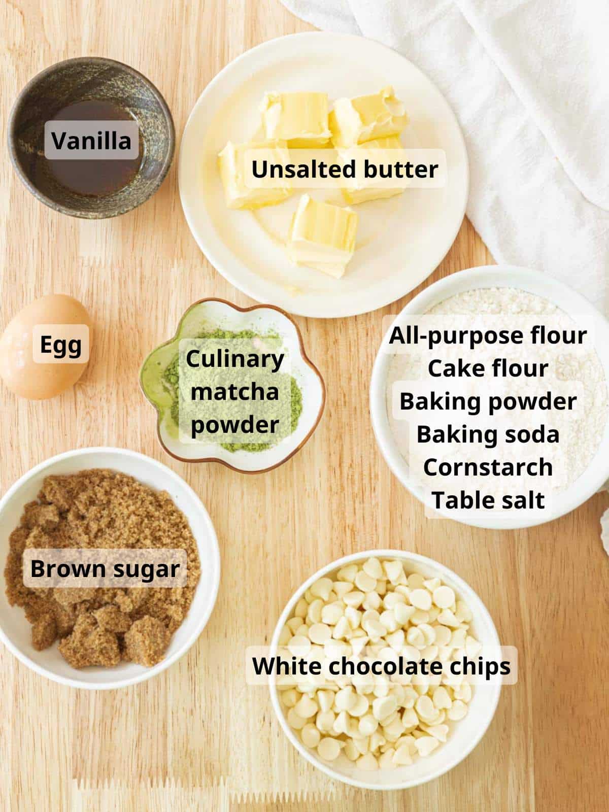 White chocolate matcha cookie ingredients laid out on a wooden table, with labels for each ingredient.