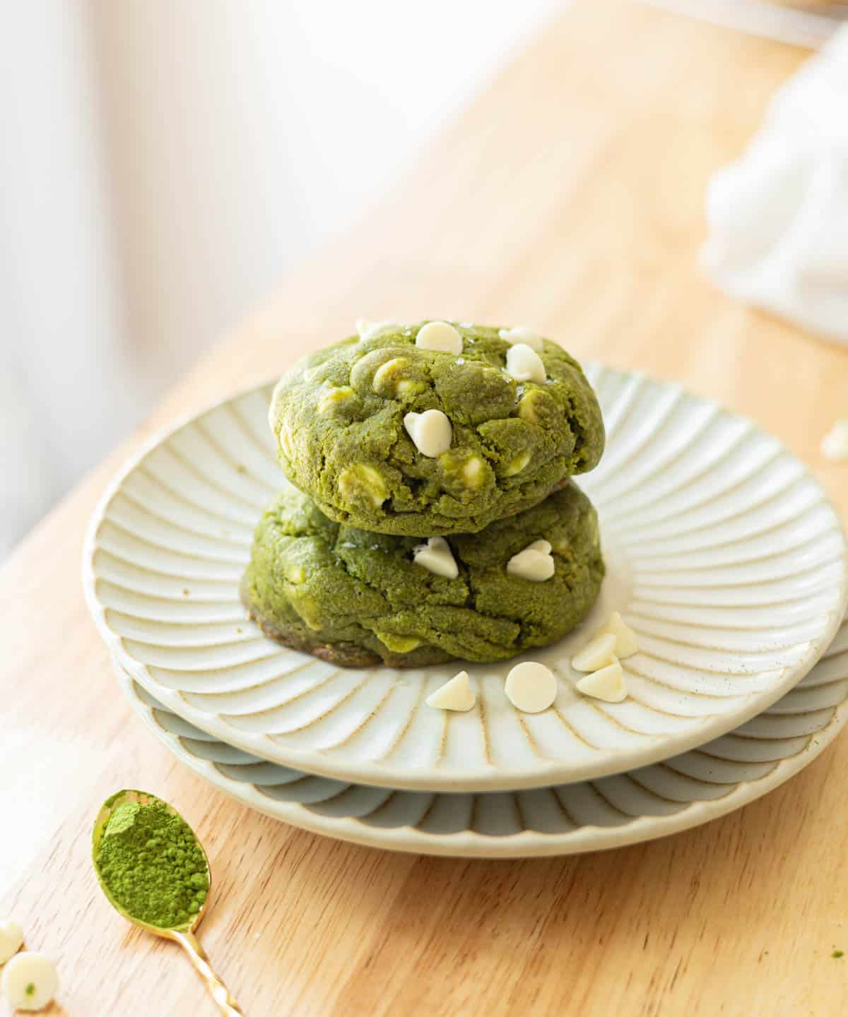 Chunky white chocolate matcha cookies on a fluted white plate on top of a wooden island.