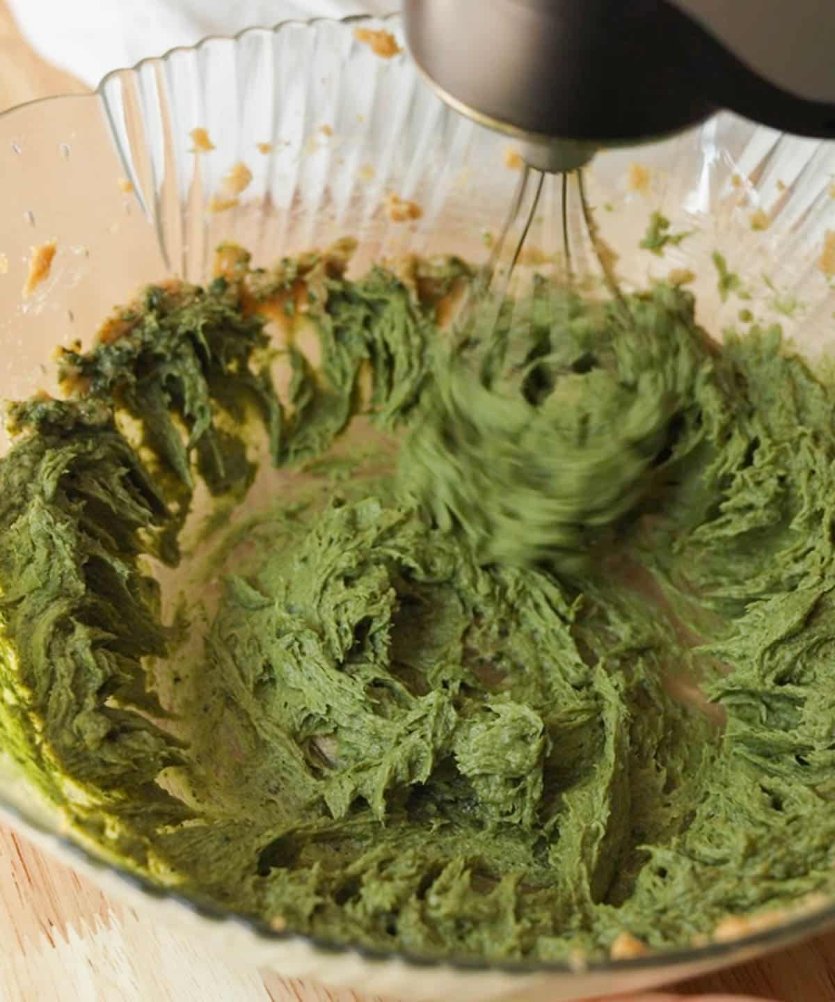 Using a hand mixer to beat green batter together in glass mixing bowl.