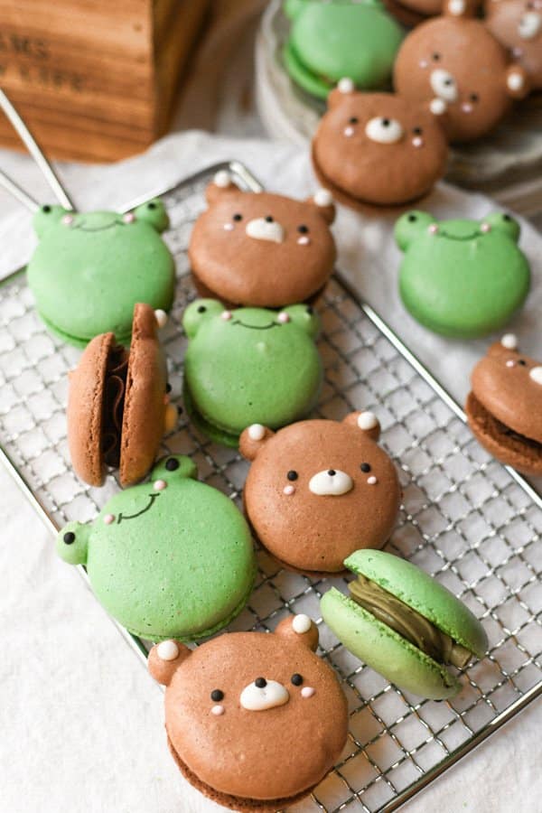 Angled photo of chocolate bear macarons and white chocolate frog macarons on a rectangular wire rack over a dining cloth 