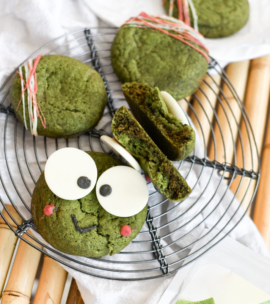 Two keroppi matcha sugar cookies (one is ripped in half, showing hte insides) on a circular wire rack, next to plain matcha sugar cookies with stripes of white + red white chocolate on them.