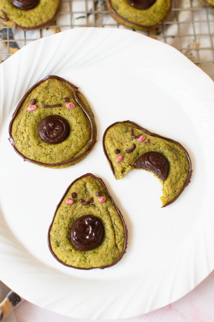 Cute avocado cookies on a white plate; one has a bite mark.