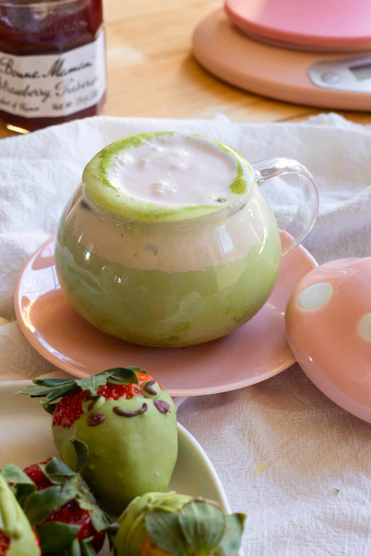 Matcha latte topped with strawberry cold foam in a cup, next to a plate with frog matcha chocolate covered strawberries.