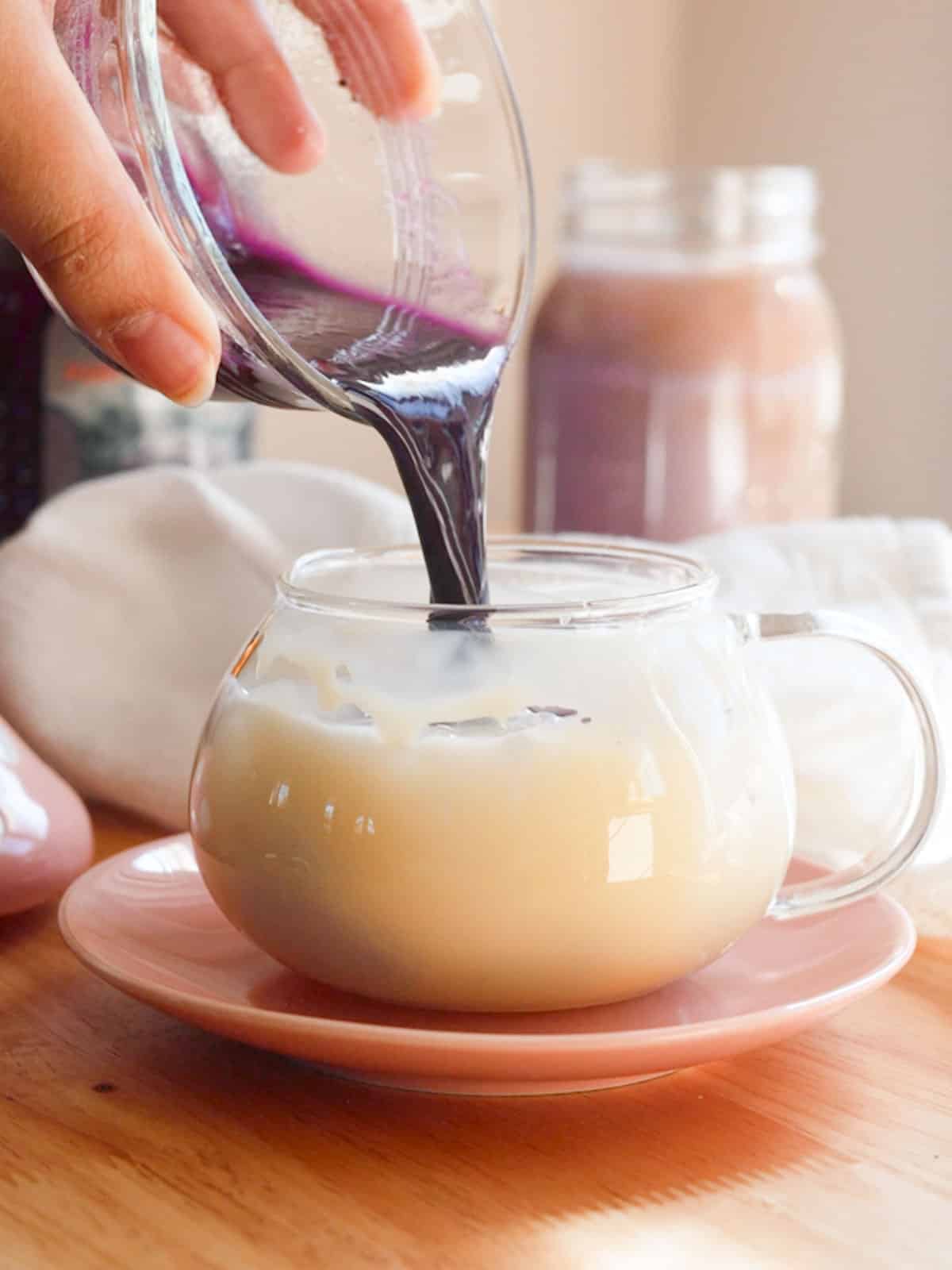 Pouring ube jam mixed with water into a glass mug with milk, ice, and condensed milk.