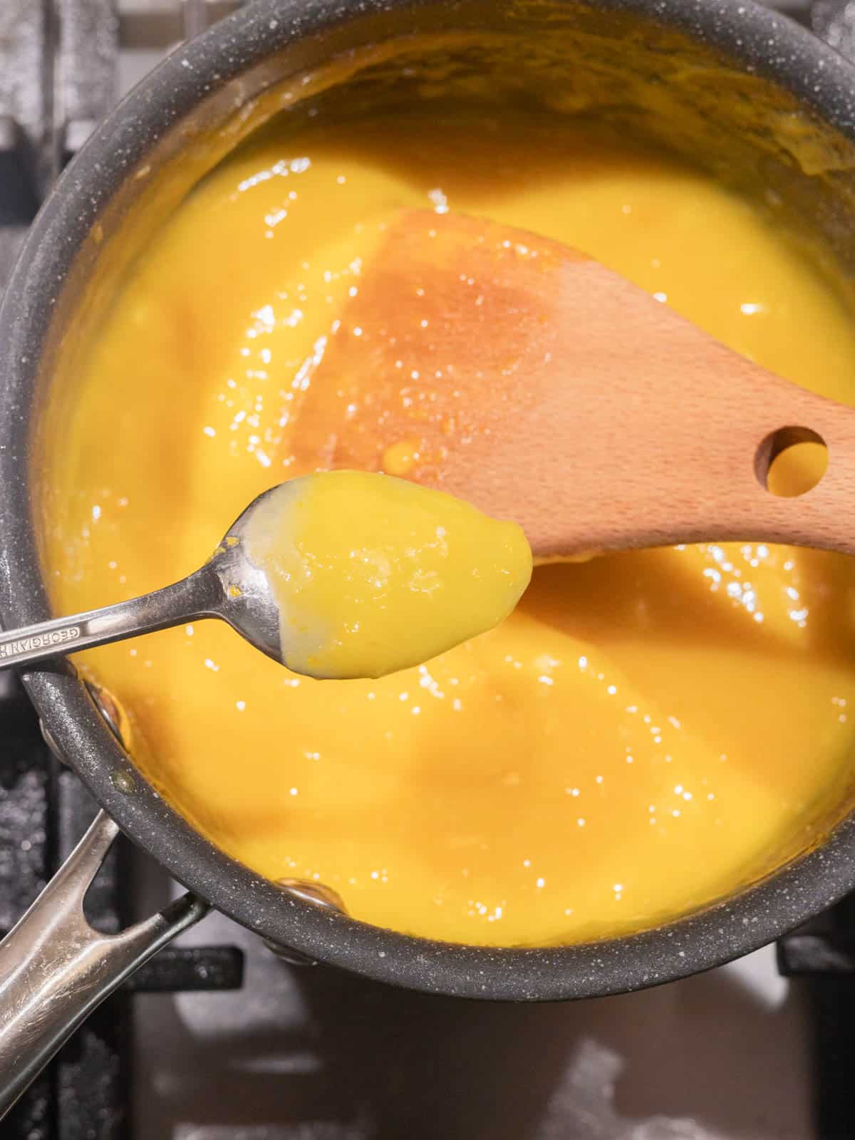 Finished lemon curd in saucepan; metal spoon has layer of lemon curd on it.