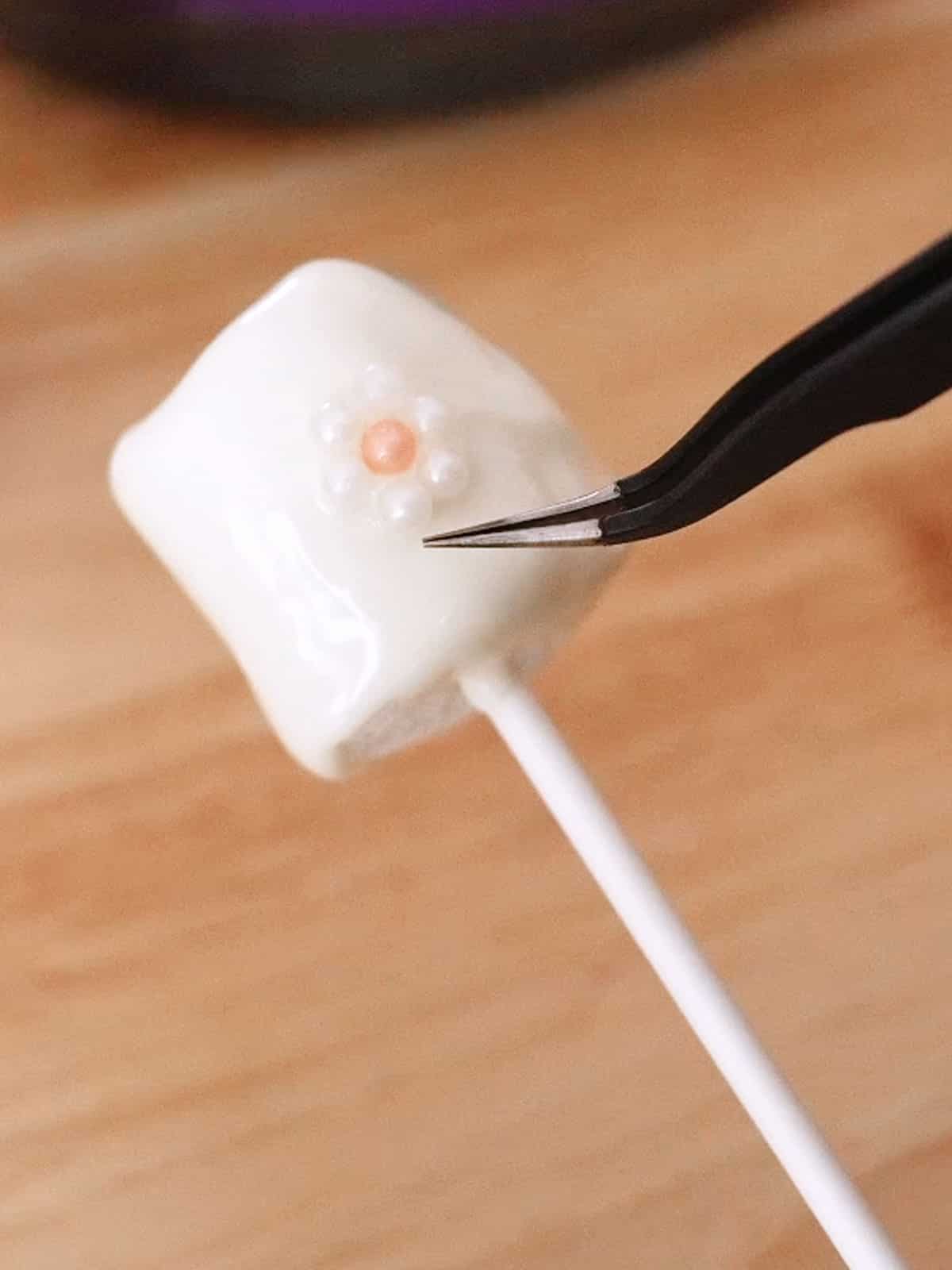 Adding sprinkles using tweezers to marshmallow pop.