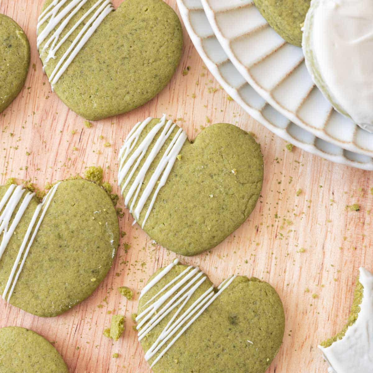 Heart-shaped matcha cut out cookies with white chocolate drizzled on top, on a light wooden backdrop.