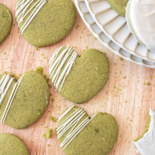 Heart-shaped matcha cut out cookies with white chocolate drizzled on top, on a light wooden backdrop.