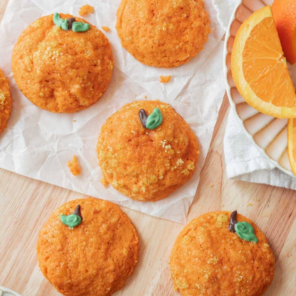 Cute orange sugar cookies decorated as oranges on a light wooden backdrop next to a cut orange fruit.