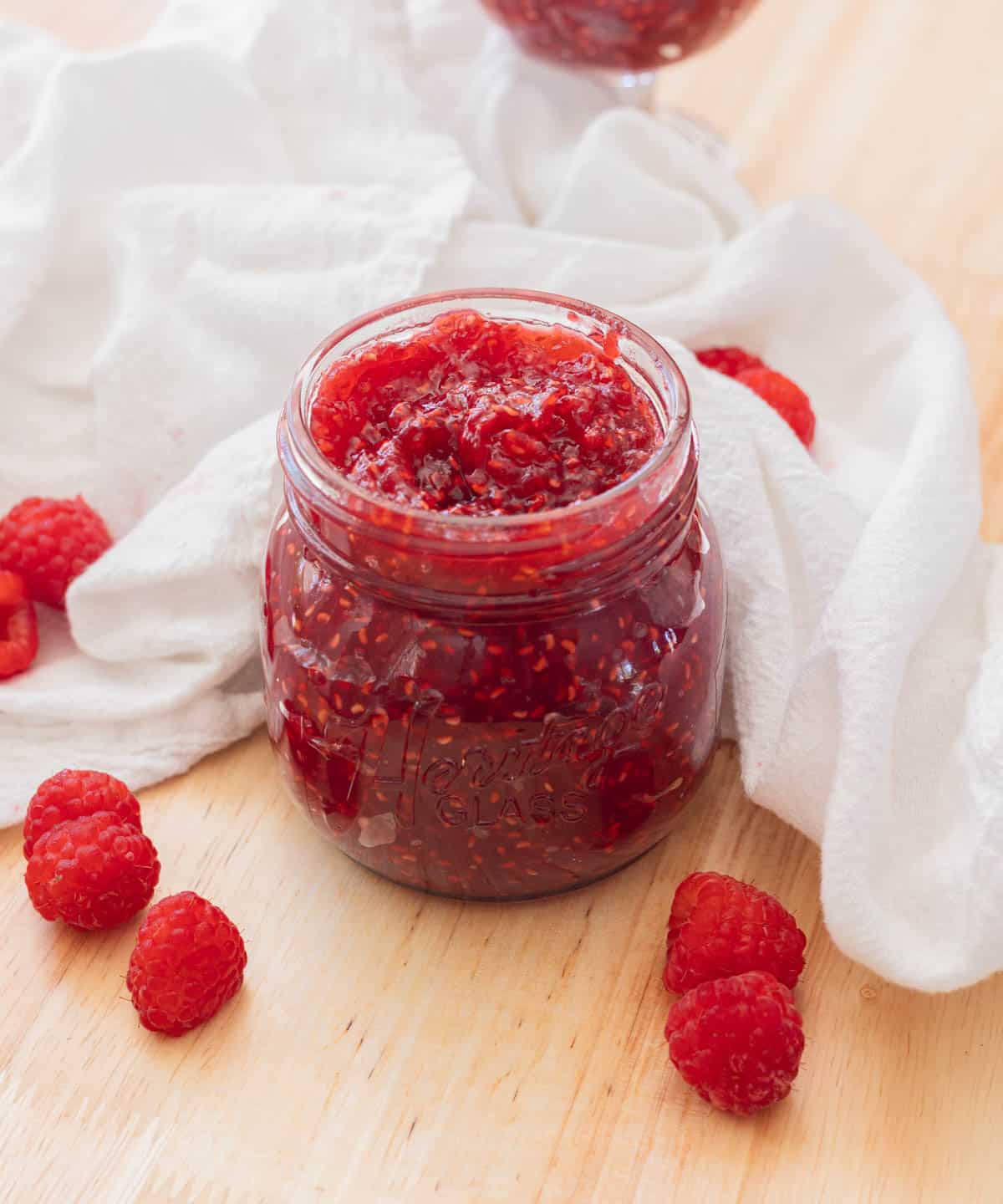 Delicious red raspberry jam in a glass jar on a wooden backdrop, with raspberries scattered around it.