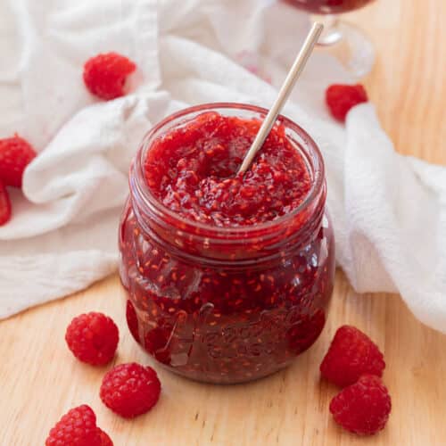 Delicious red raspberry jam in a glass jar on a wooden backdrop, with raspberries scattered around it.