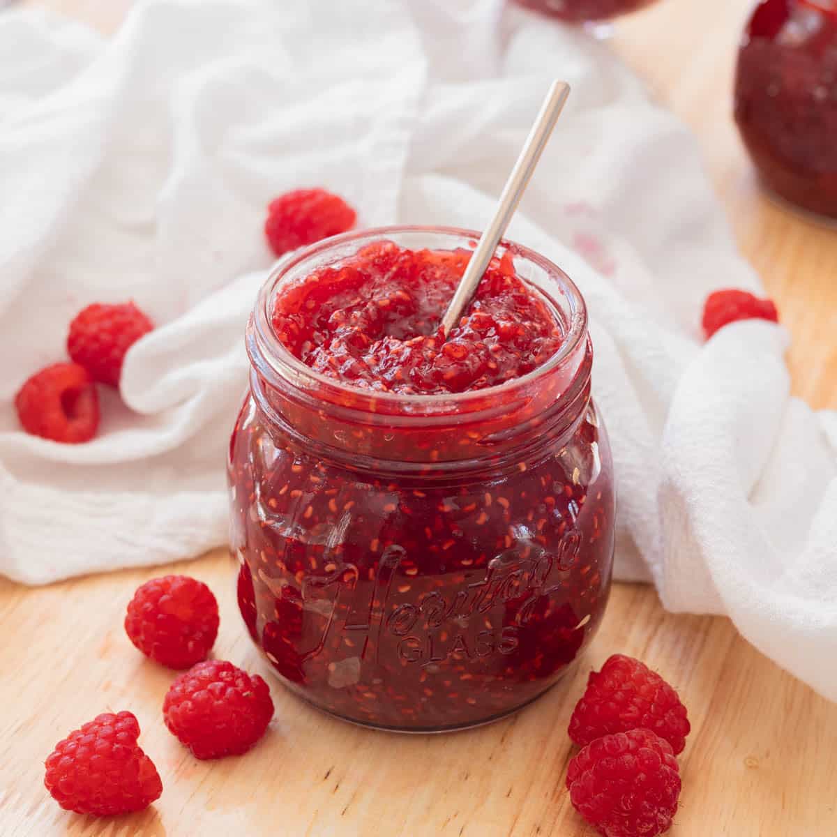 Delicious red raspberry jam in a glass jar on a wooden backdrop, with raspberries scattered around it.