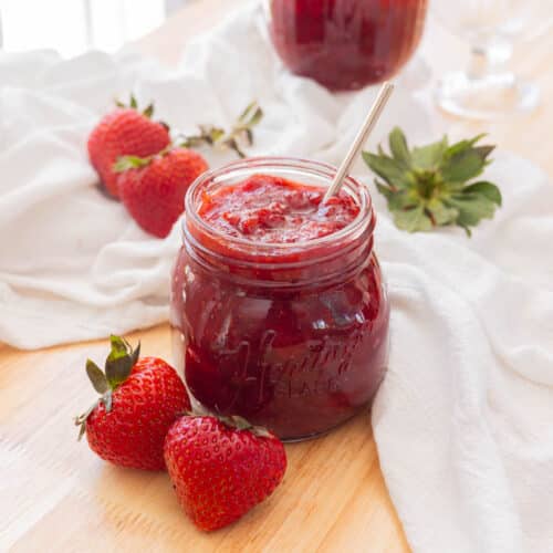 Delicious red strawberry jam in a glass jar on a wooden backdrop, with two strawberries in front of it.