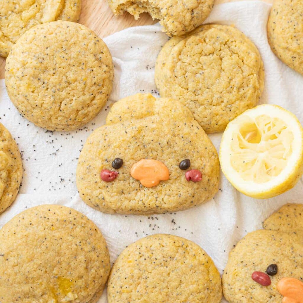 Cute duck-shaped lemon curd cookie on a white linen, surrounded by regular lemon curd cookies, half a squeezed lemon, and poppy seeds.