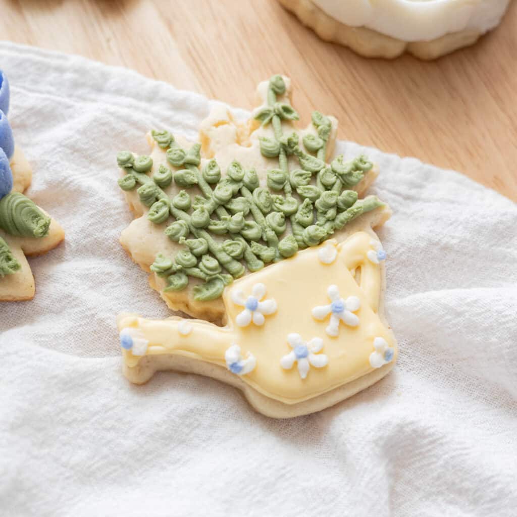 Yellow watering can sugar cookie with green plants sticking out from the top.