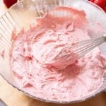 Pink strawberry whipped cream frosting in a glass bowl with a whisk, which has stiff peaks.