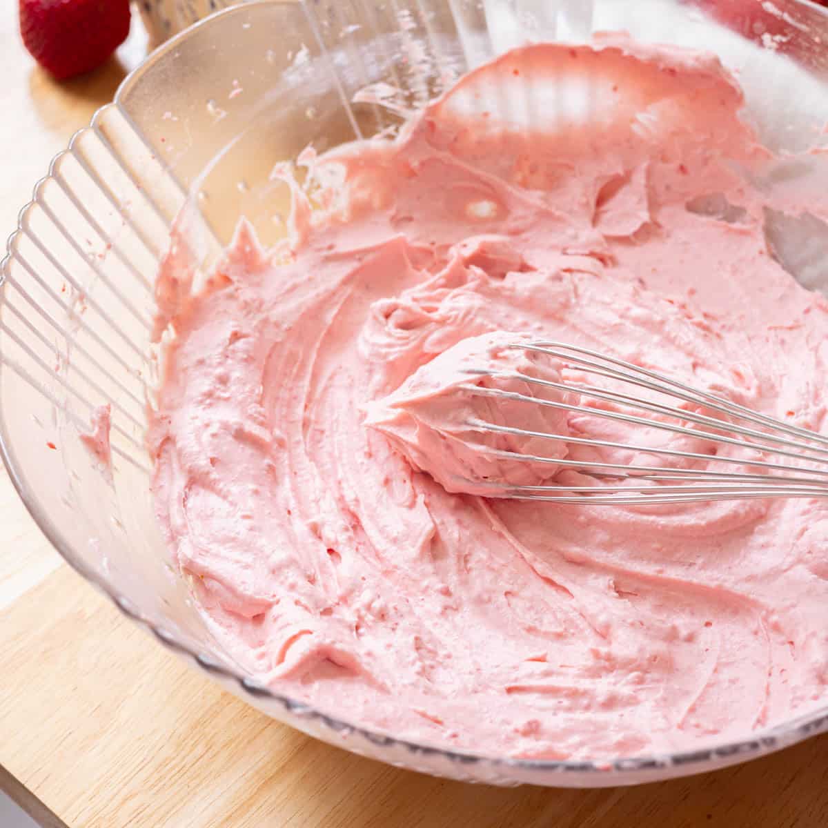 Pink strawberry whipped cream frosting in a glass bowl with a whisk, which has stiff peaks.