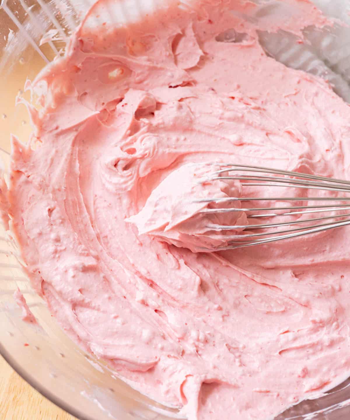 Stiff peak strawberry whipped cream frosting on a whisk on top of a bowl with the rest of the pink frosting.