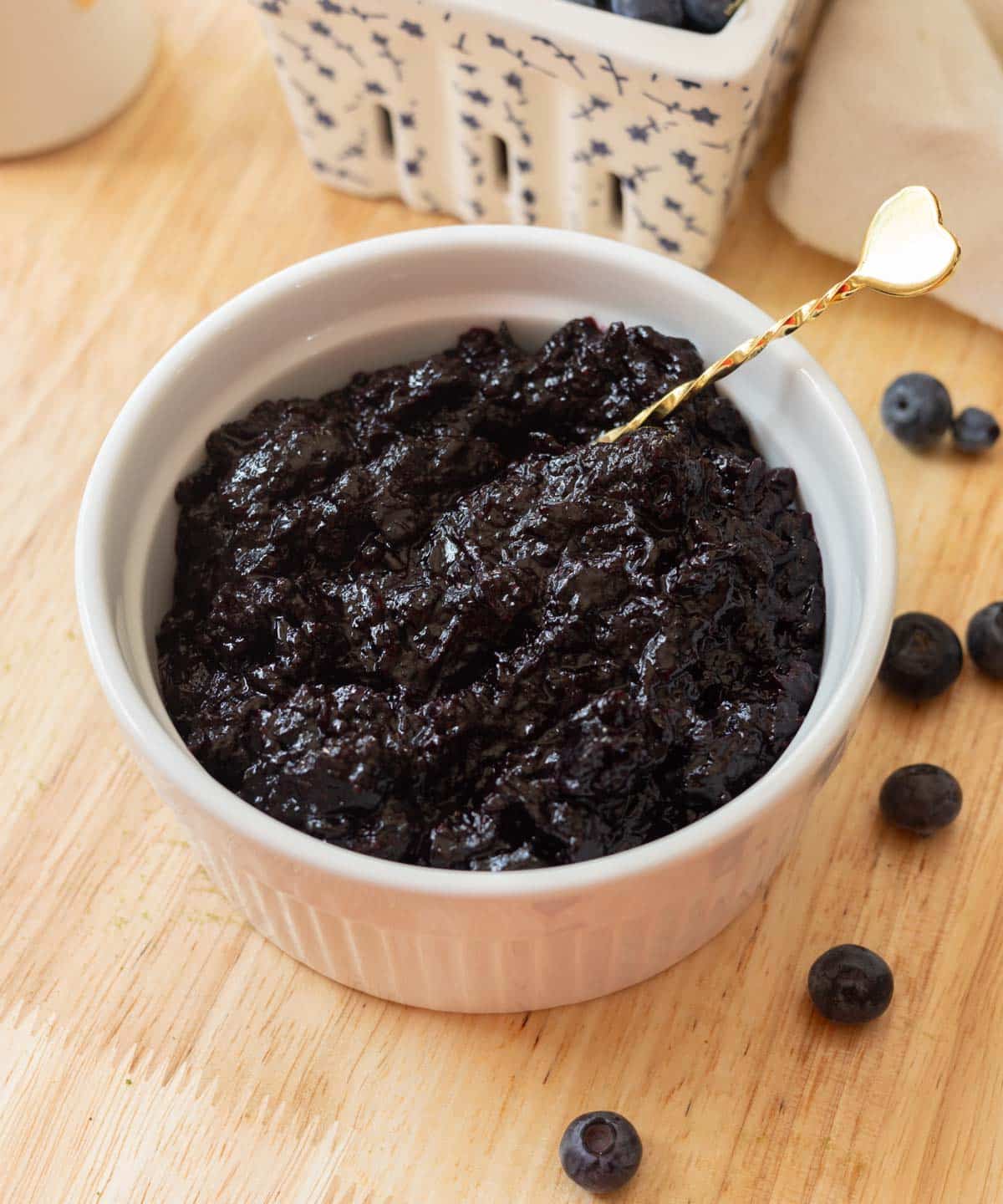 Blueberry jam in a white ramekin on a wooden table, surrounded by a few blueberries.