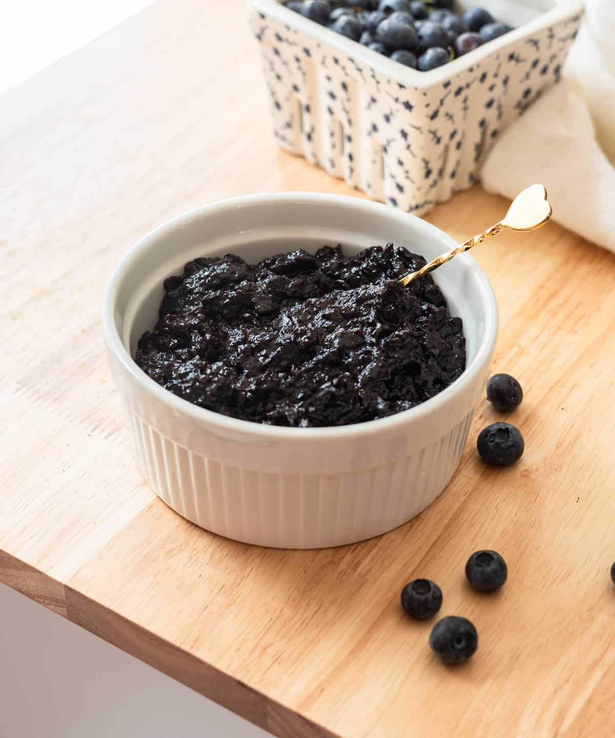 Blueberry jam in a white ramekin on a wooden table, surrounded by a few blueberries.