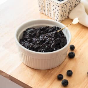 Blueberry jam in a white ramekin on a wooden table, surrounded by a few blueberries.