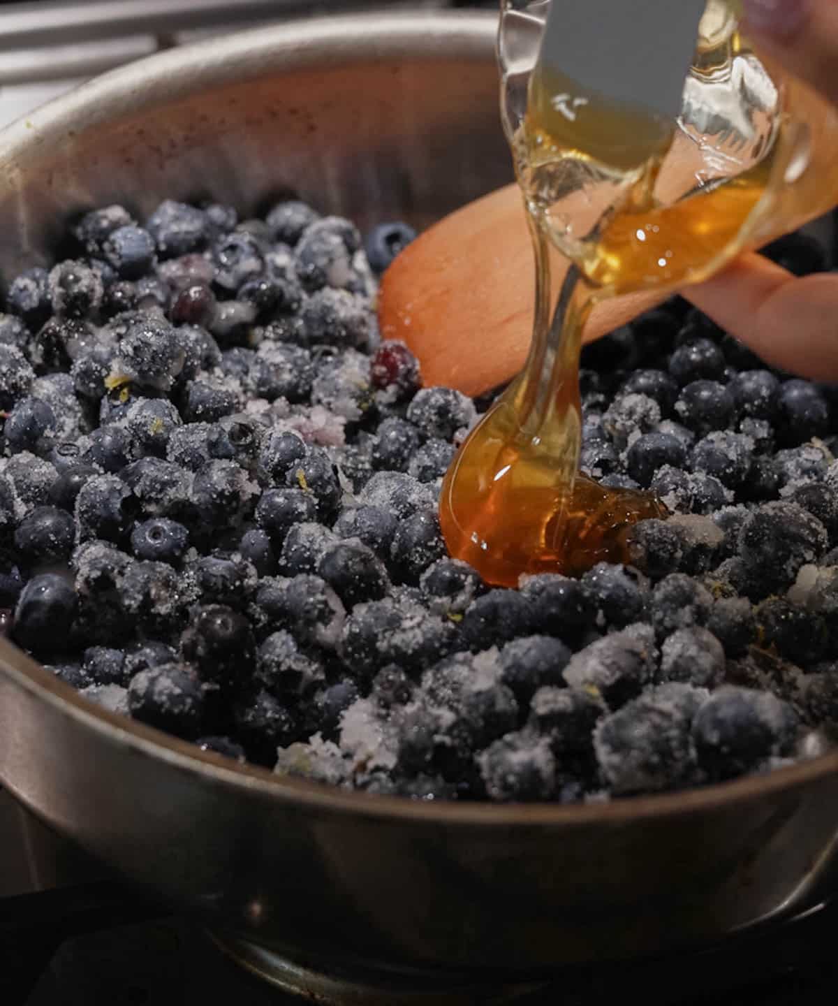 Pouring honey into a large saucepan with blueberries, granulated sugar, lemon juice, and lemon zest.