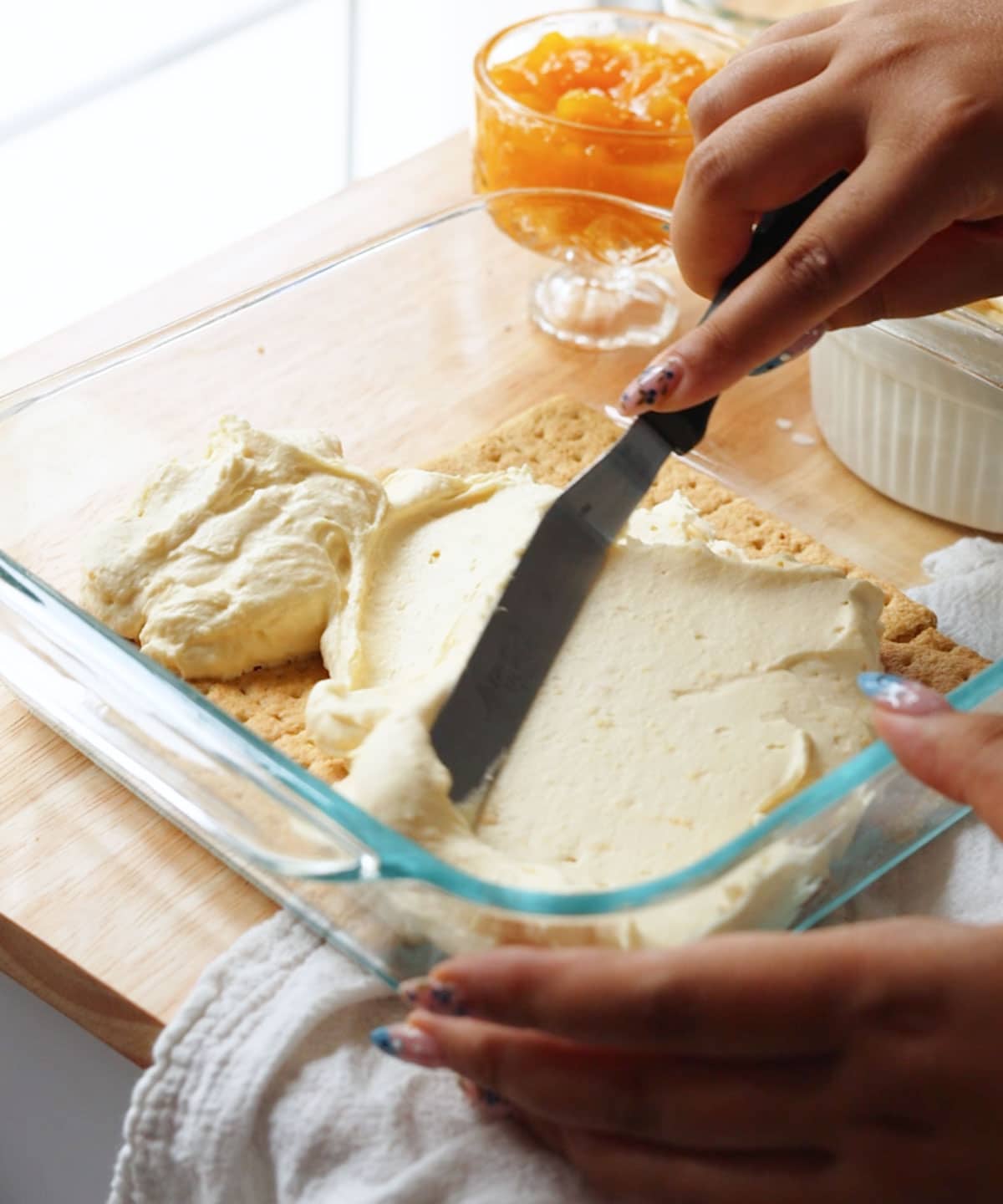 Using an offset spatula to spread mango whipped cream over a layer of graham crackers in a glass baking dish.