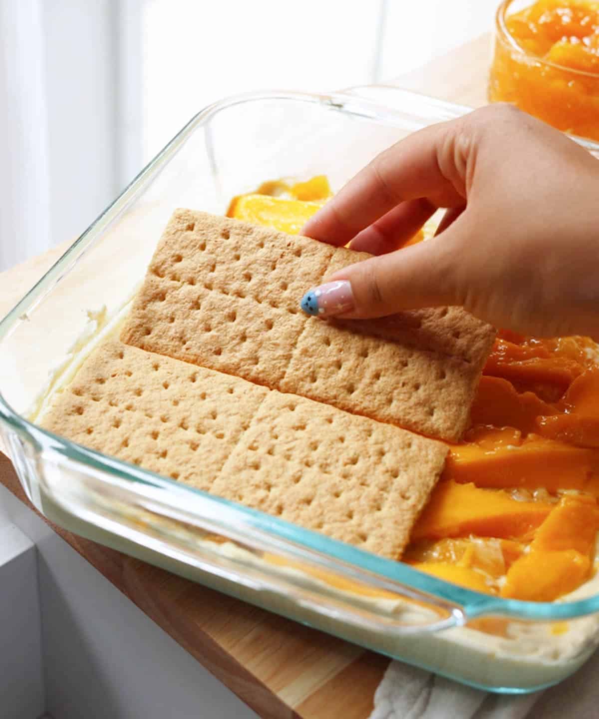 Adding a layer of graham crackers on top of sliced mangoes.