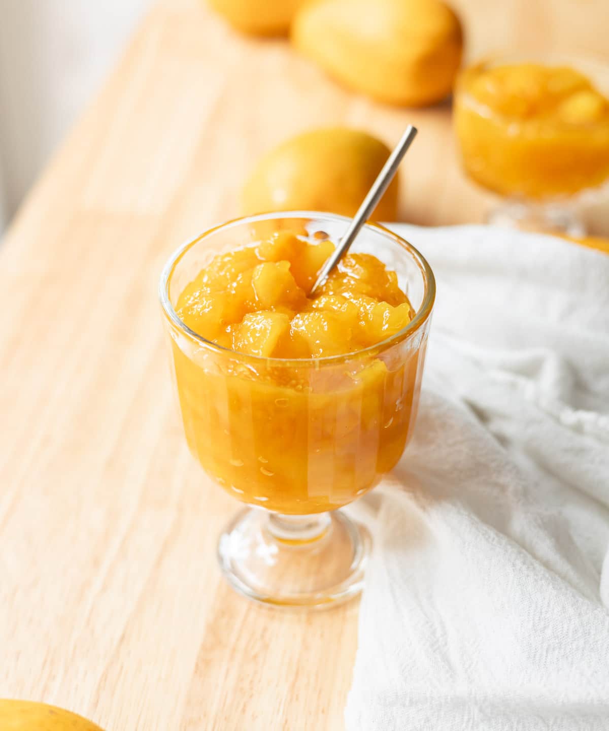 Vibrant orange yellow mango jam in a glass serving cup on a wooden table, surrounded by a few mangoes.