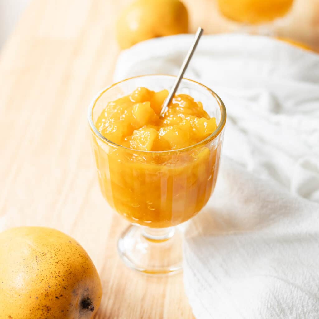 Vibrant orange yellow mango jam in a glass serving cup on a wooden table, surrounded by a few mangoes.