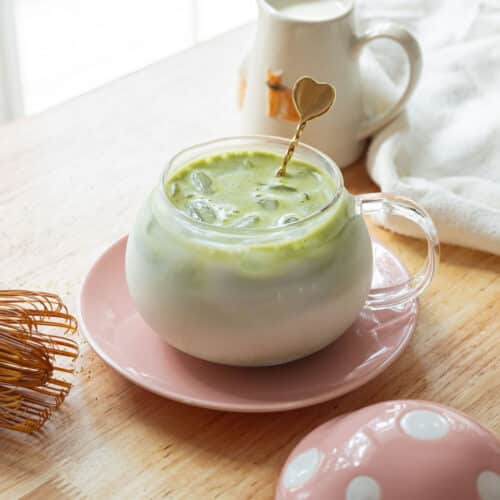 Iced matcha latte in a glass cup on a pink dish, over a light-colored wooden table.