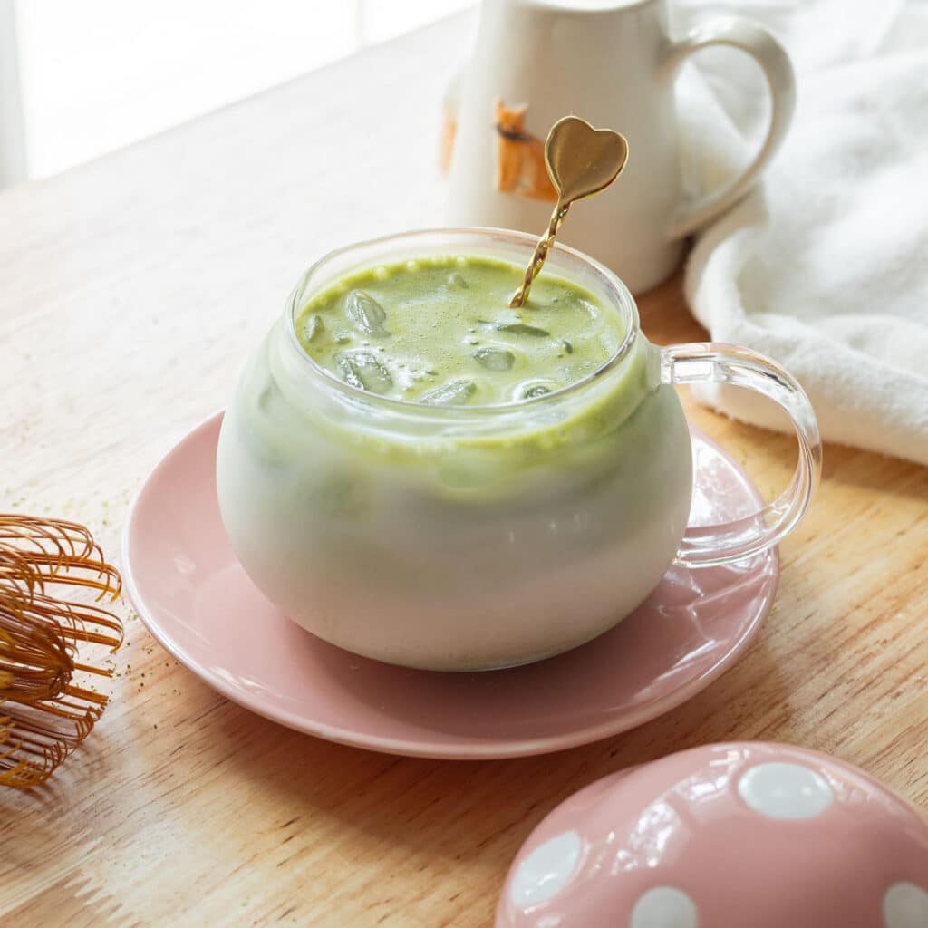 Iced matcha latte in a glass cup on a pink dish, over a light-colored wooden table.