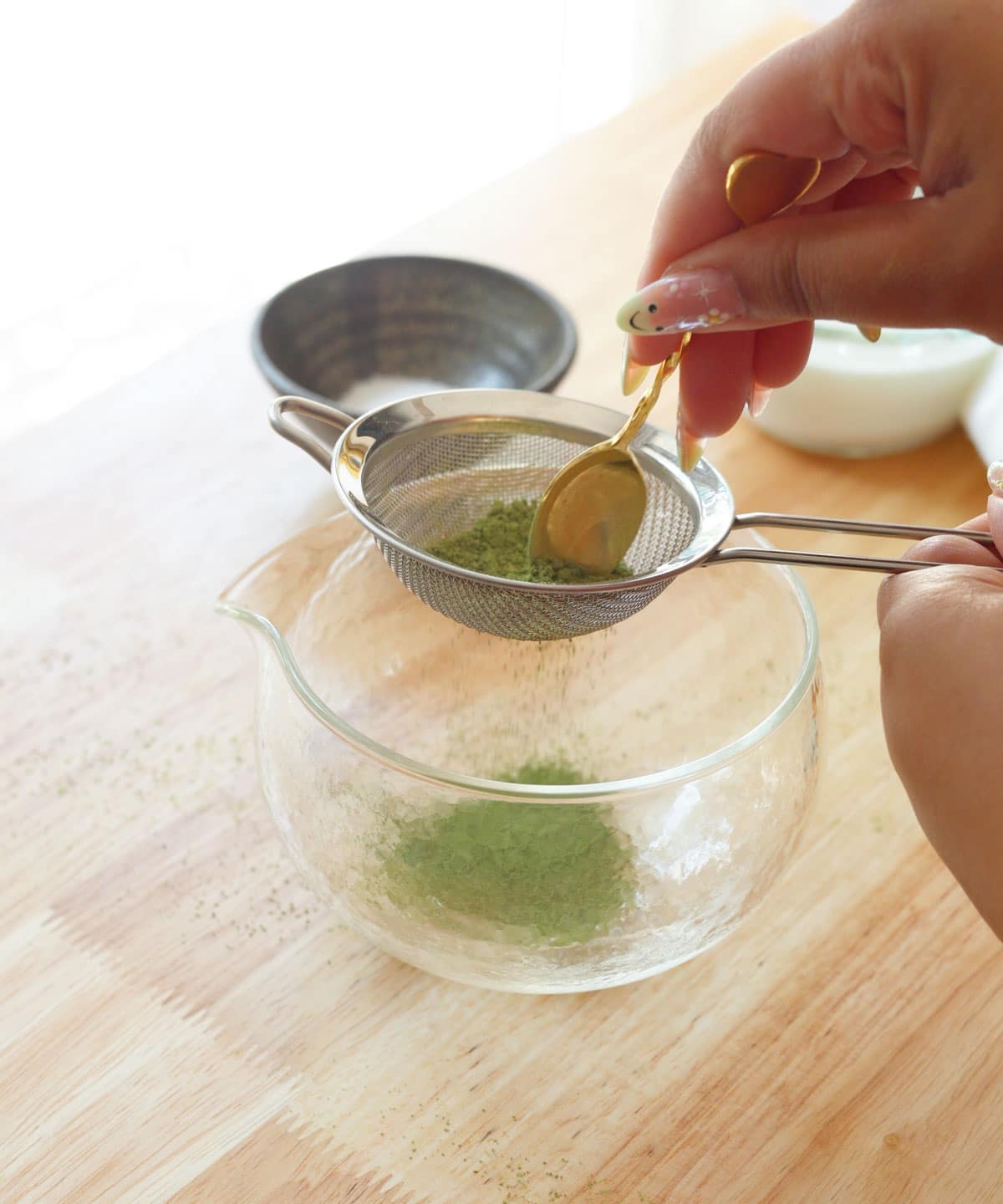 Using a small sifter to sift matcha powder into a glass matcha bowl.