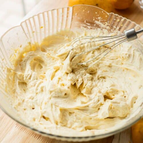 Stiff peak mango whipped cream frosting on a whisk on top of a bowl with the rest of the naturally yellow orange frosting.