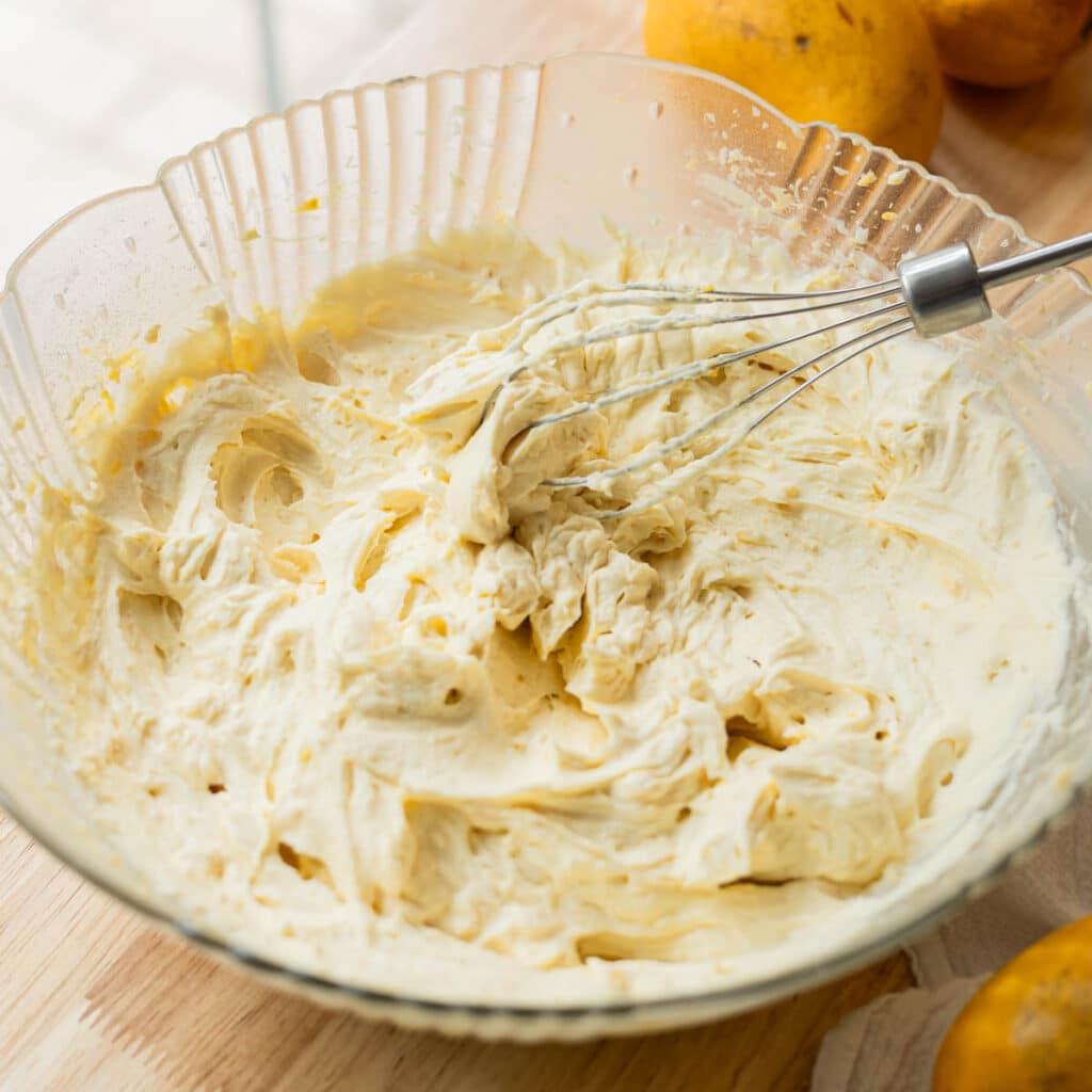 Stiff peak mango whipped cream frosting on a whisk on top of a bowl with the rest of the naturally yellow orange frosting.