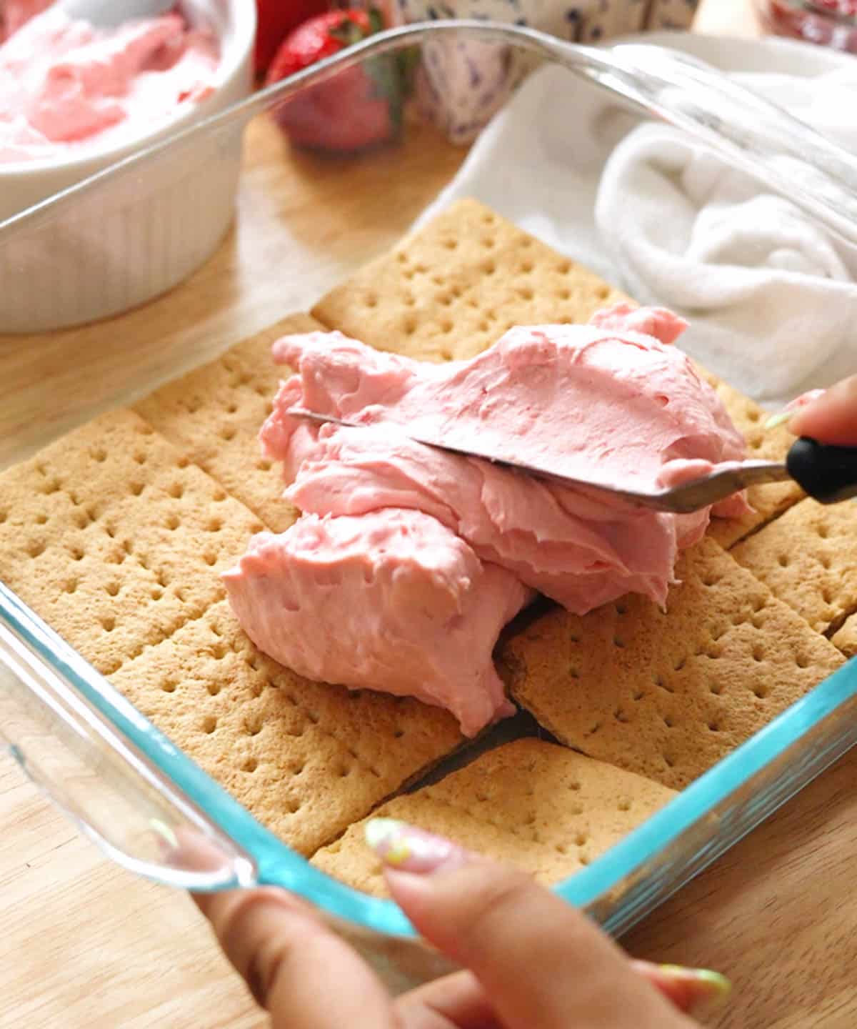 Using an offset spatula to spread strawberry whipped cream over a layer of graham crackers in a glass baking dish.