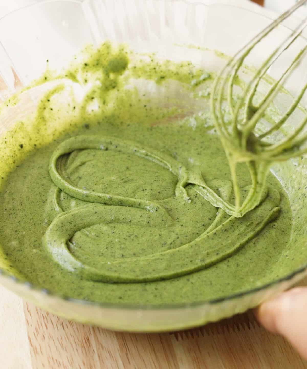 Glass mixing bowl with green batter in it; a heart is drawn on top using batter by using the whisk.