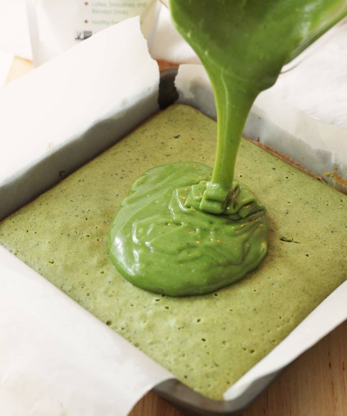 Pouring green matcha ganache on top of mochi brownies in pan.