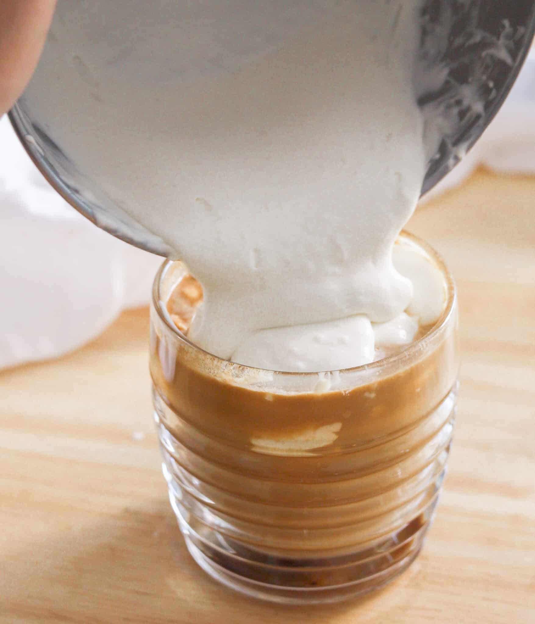 Pouring cheese foam on top of latte in glass cup.