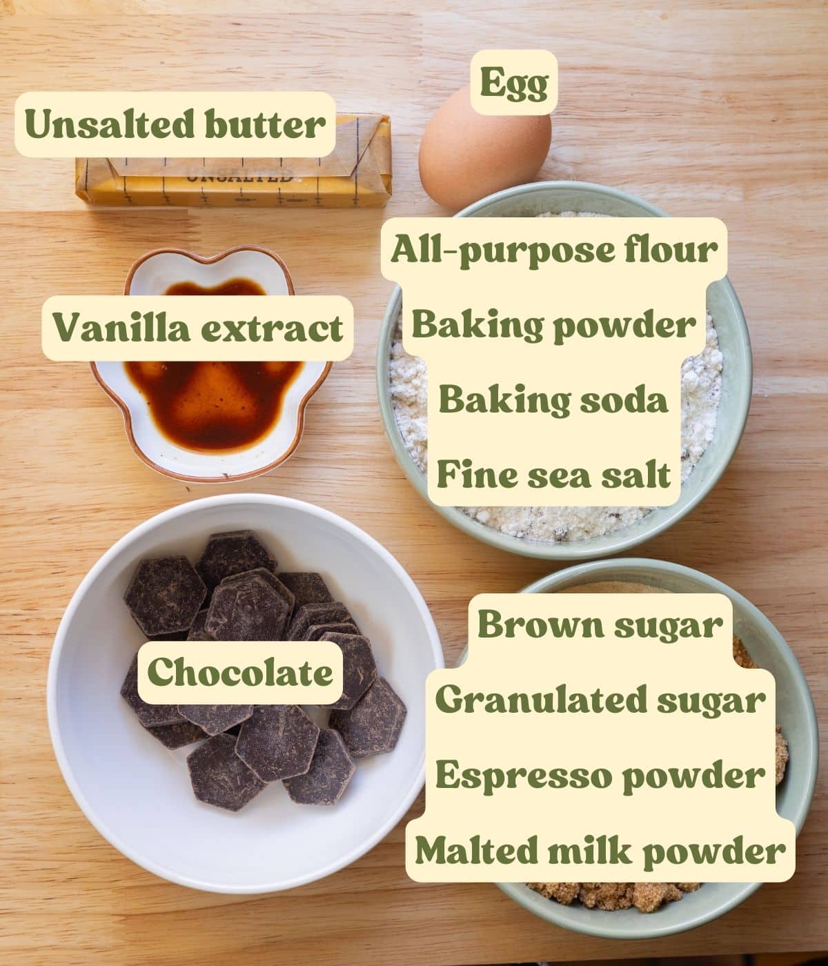 Malted milk chocolate chip cookie ingredients laid out on a wooden table, with labels for each ingredient.