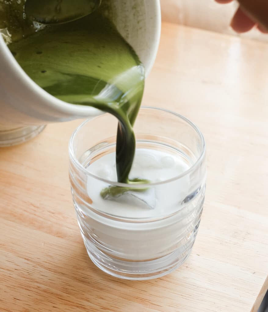 Pouring matcha mixture into a cup of oat milk.